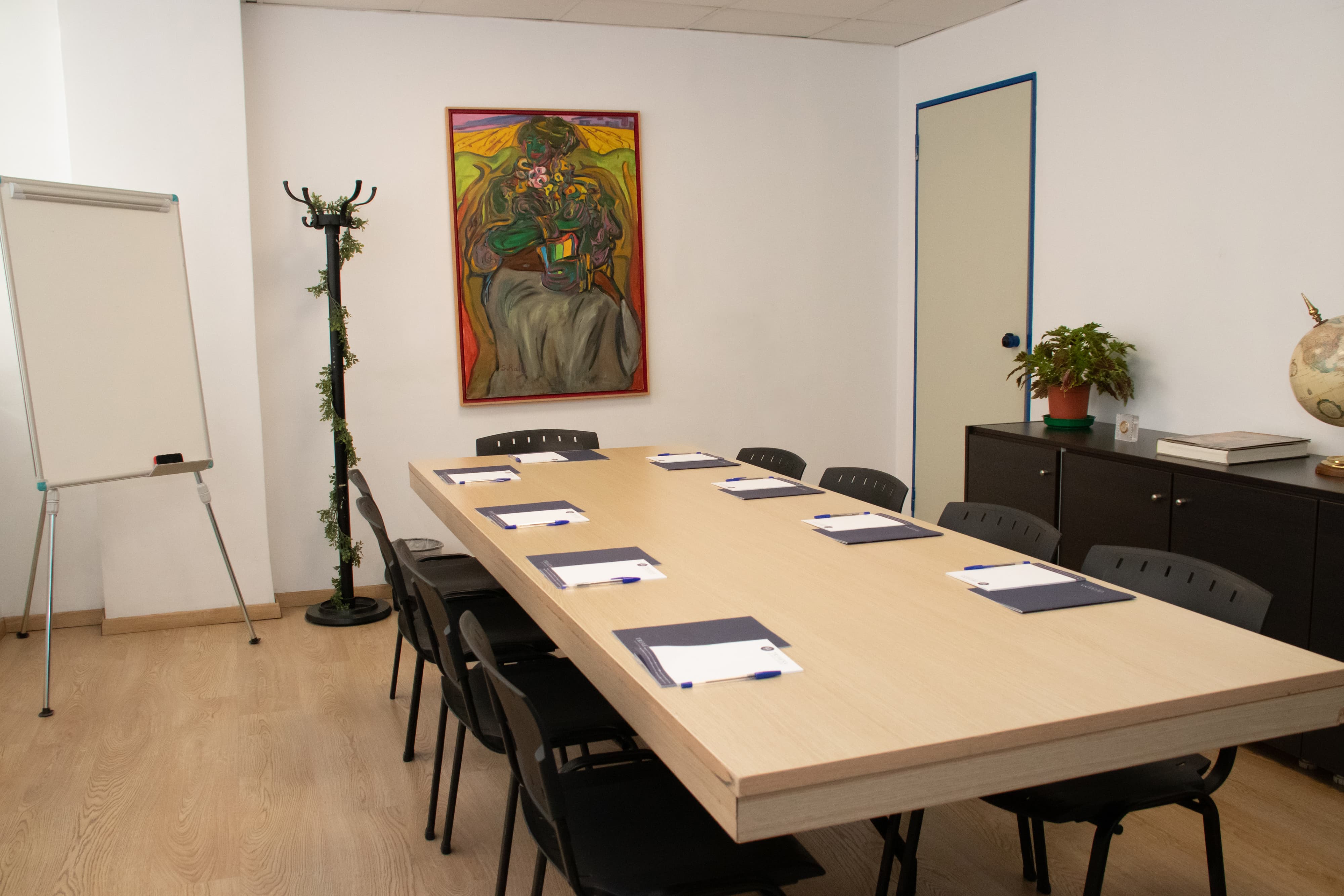 Meeting room with a rectangular wooden table surrounded by black chairs, notepads and pens on the table, a colorful painting on the wall and a flipchart stand.