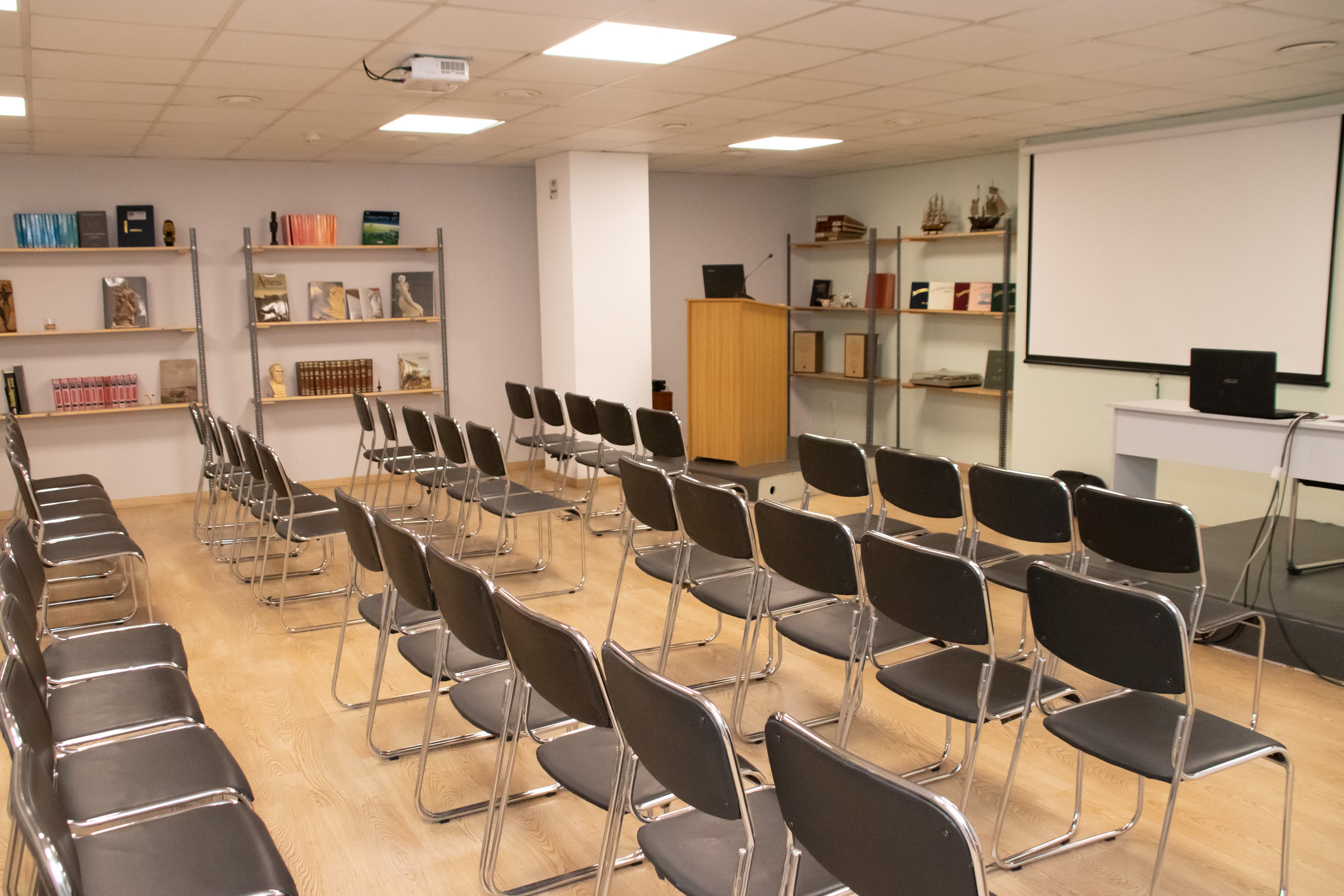 Theater-style seating arrangement with rows of chairs facing a screen, bookshelves on the side, and a small desk in front.