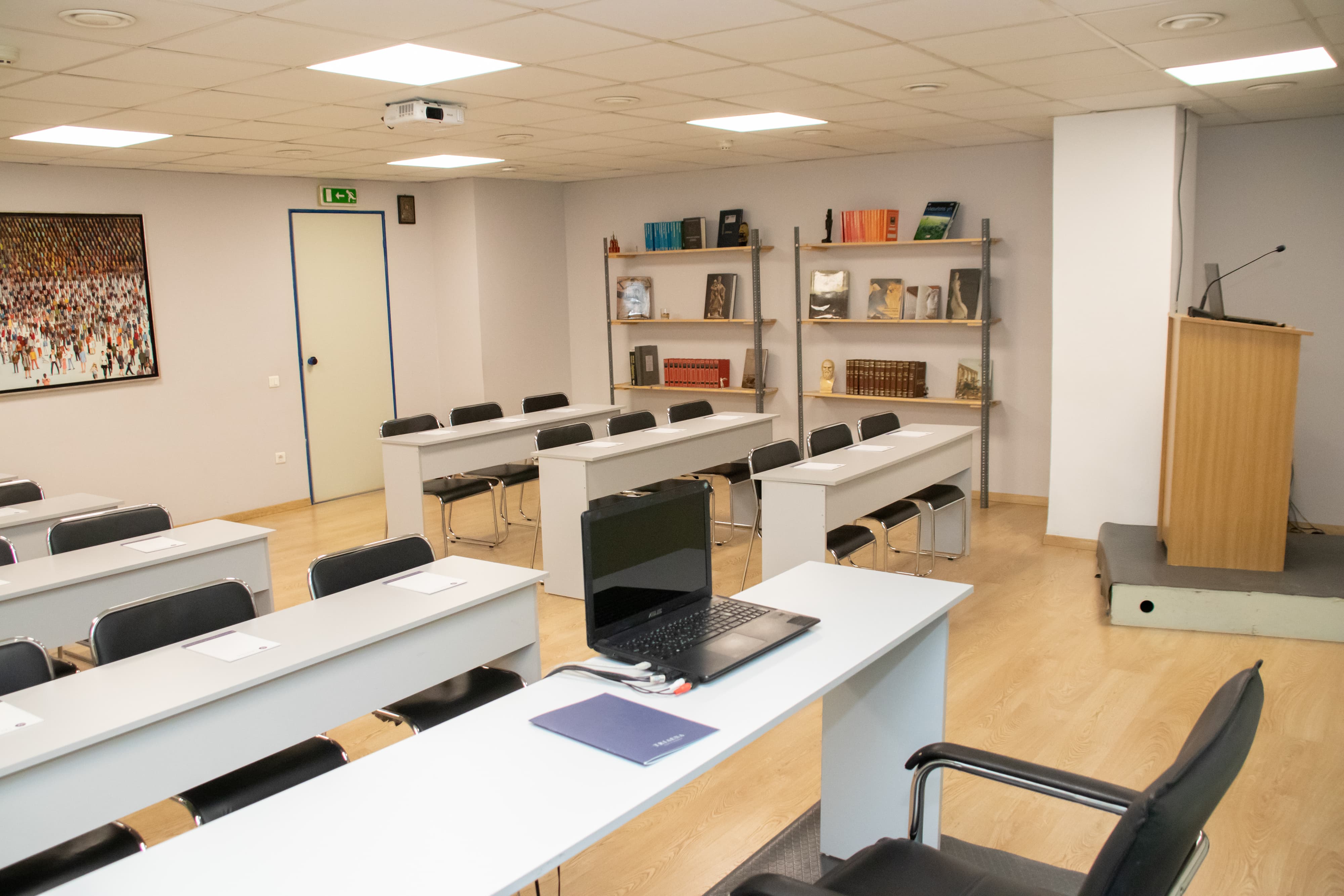 Classroom-style meeting room with rows of white tables and chairs facing forward, bookshelves on the wall, and a wooden podium in the corner.