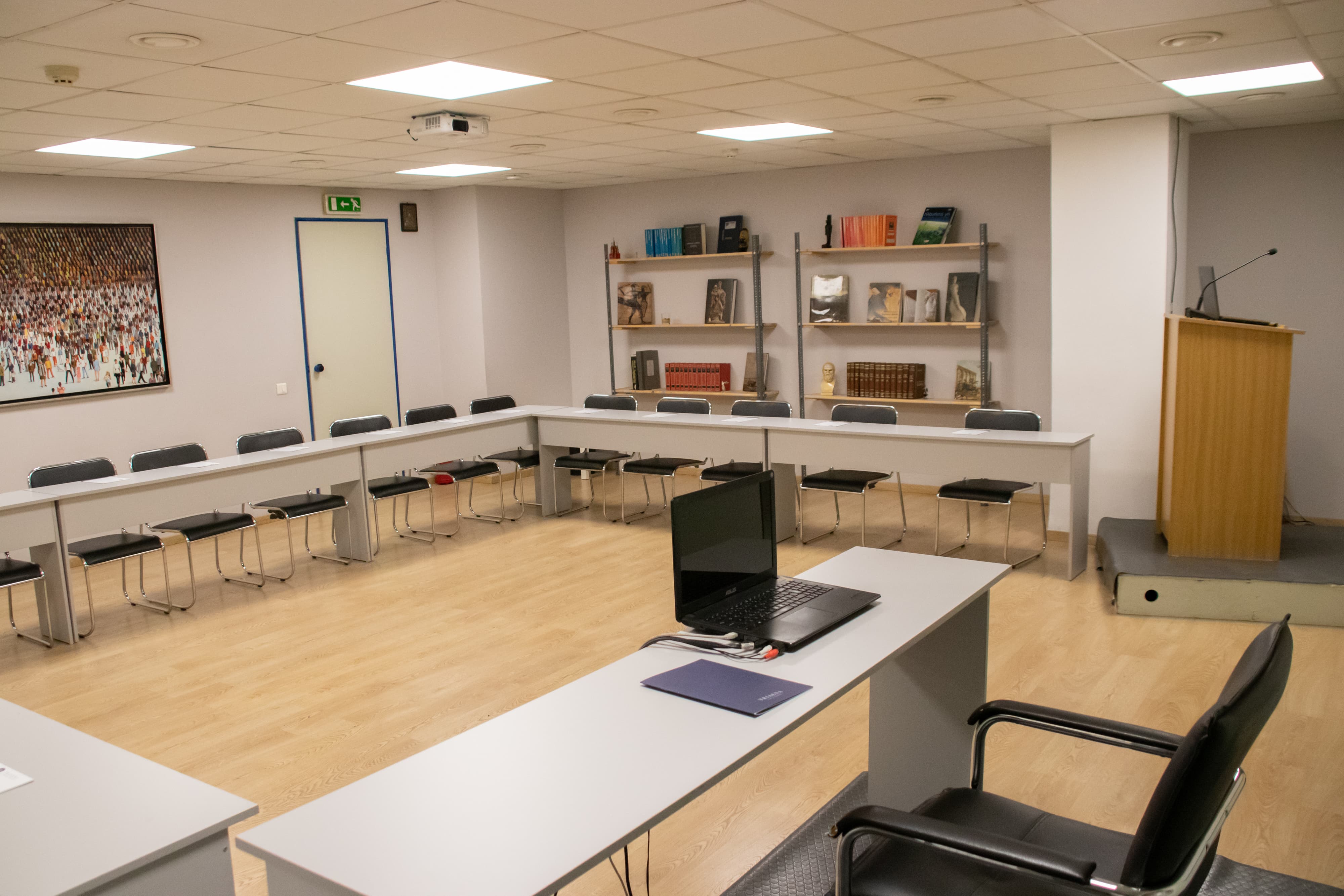 Meeting room with square table arrangement surrounded by chairs, a wooden podium, bookshelves, and framed artwork on the wall.