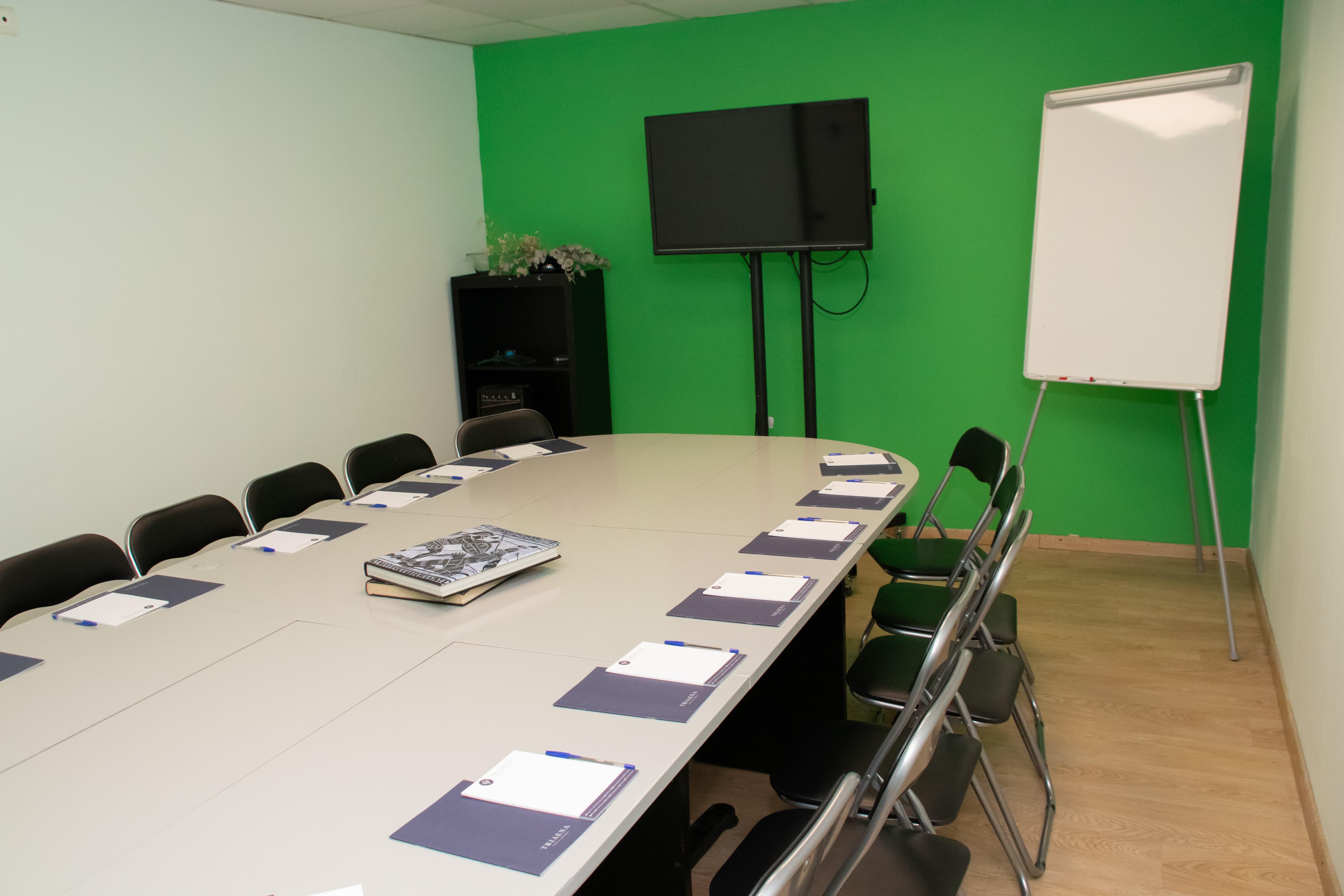 A meeting room with a green-painted wall and an oval-shaped table surrounded by chairs. A TV and a standing flipchart are placed in front of the green wall for presentations.