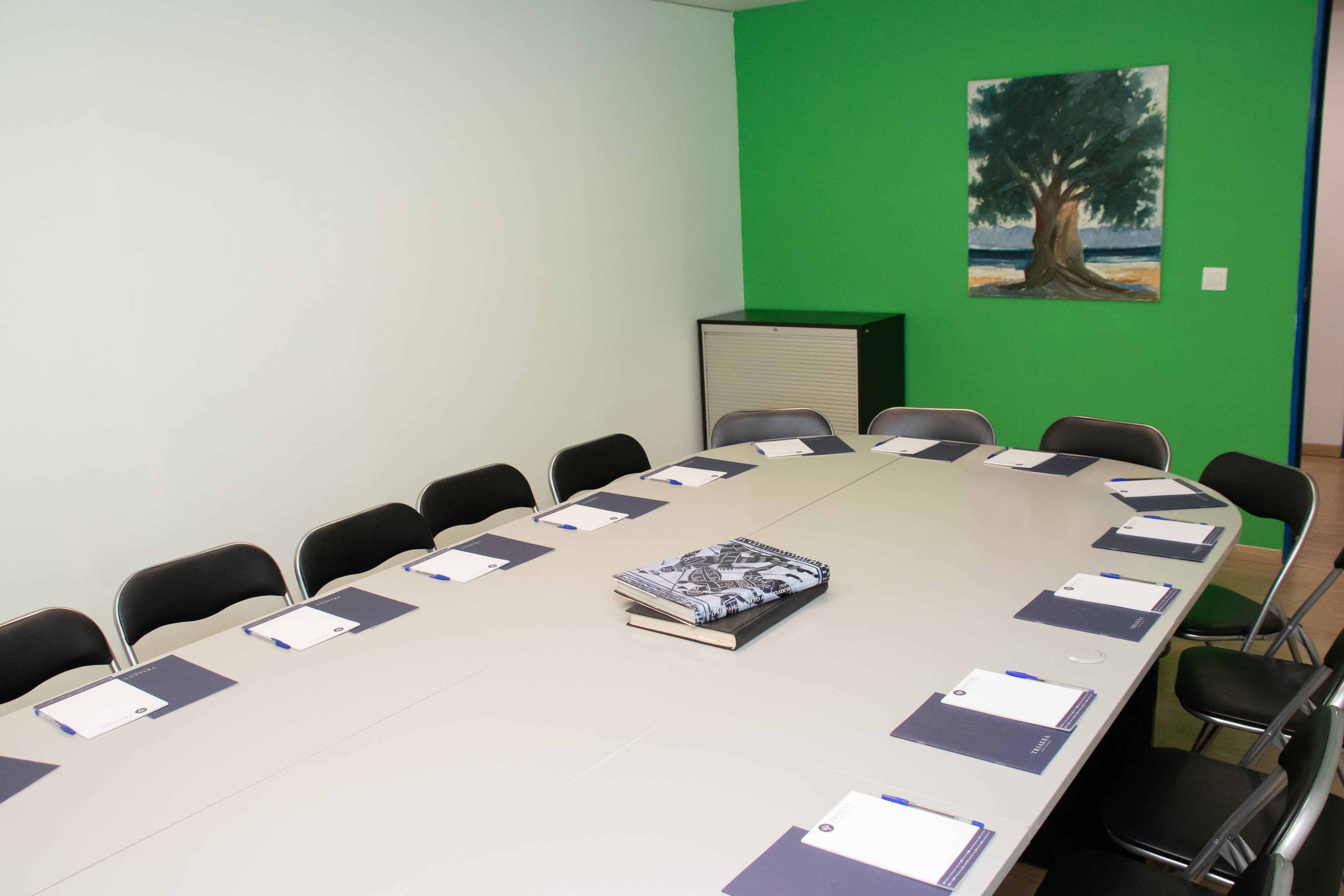 A meeting room with a green-painted wall and an oval-shaped table surrounded by chairs, and a picture of a tree hanging on the wall.