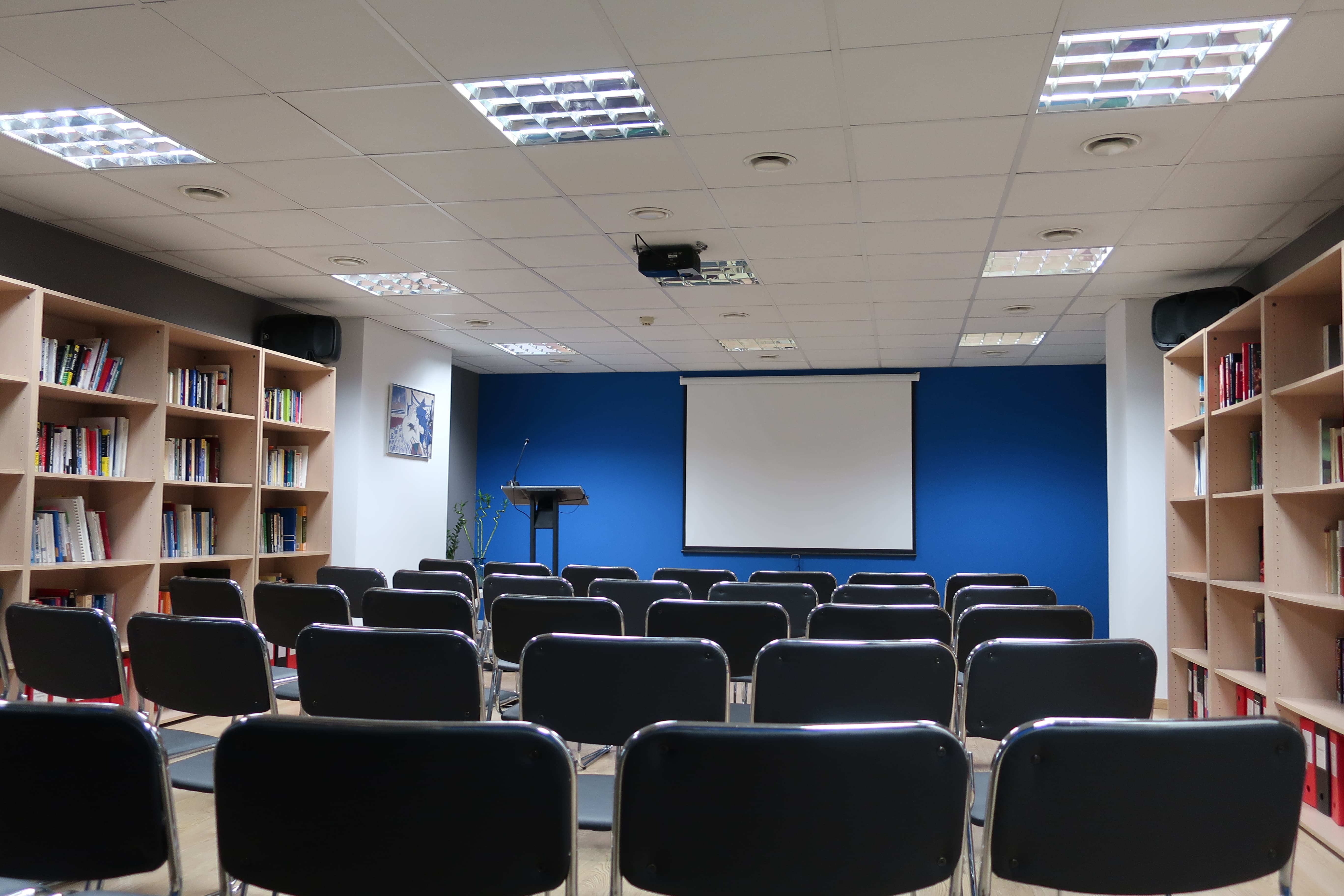 Conference room with theater-style chair arrangement, podium and screen at the front, and bookshelves on both sides.
