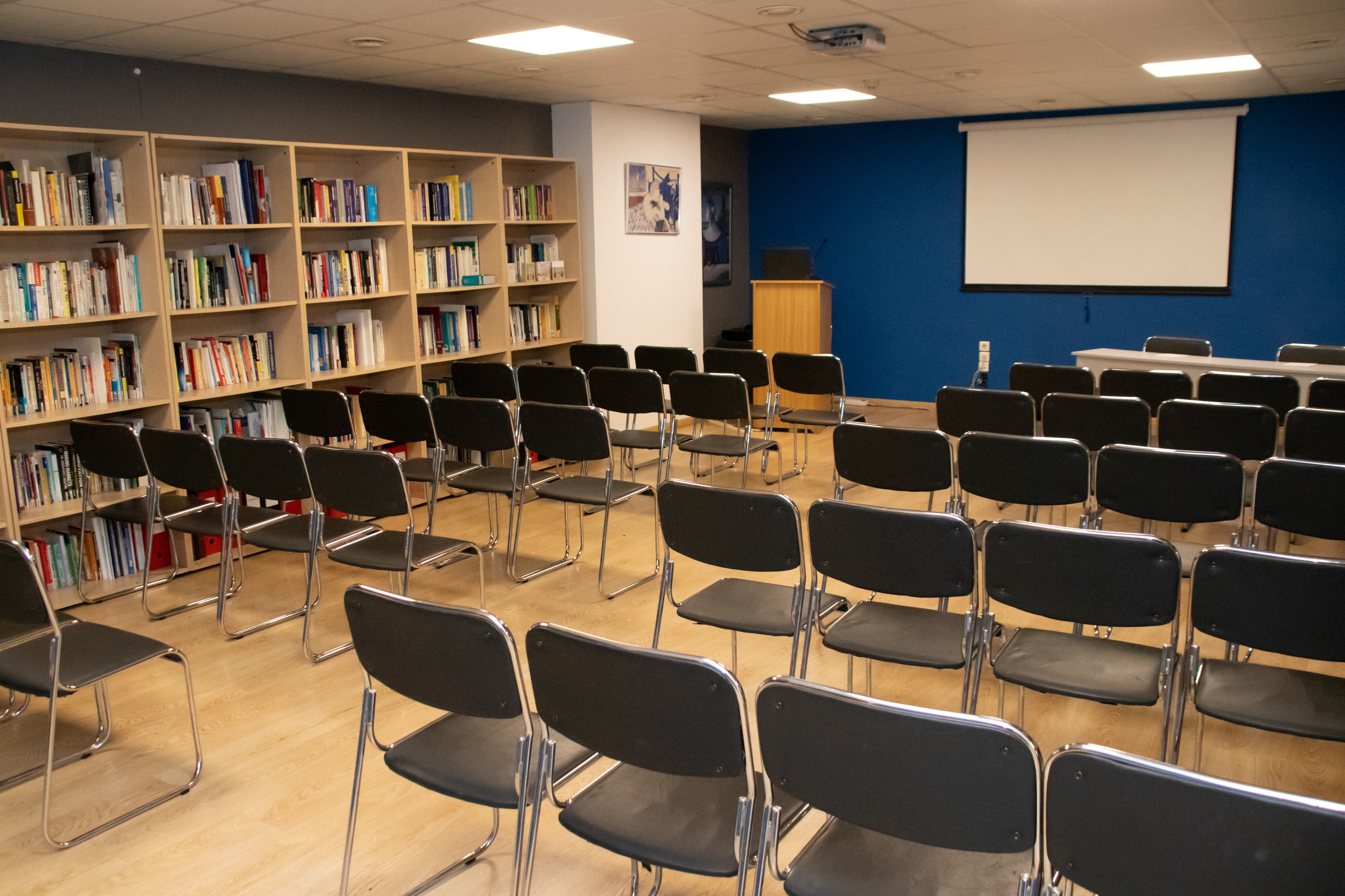 Theater-style meeting room with multiple rows of chairs facing a projection screen and bookshelf-lined walls.