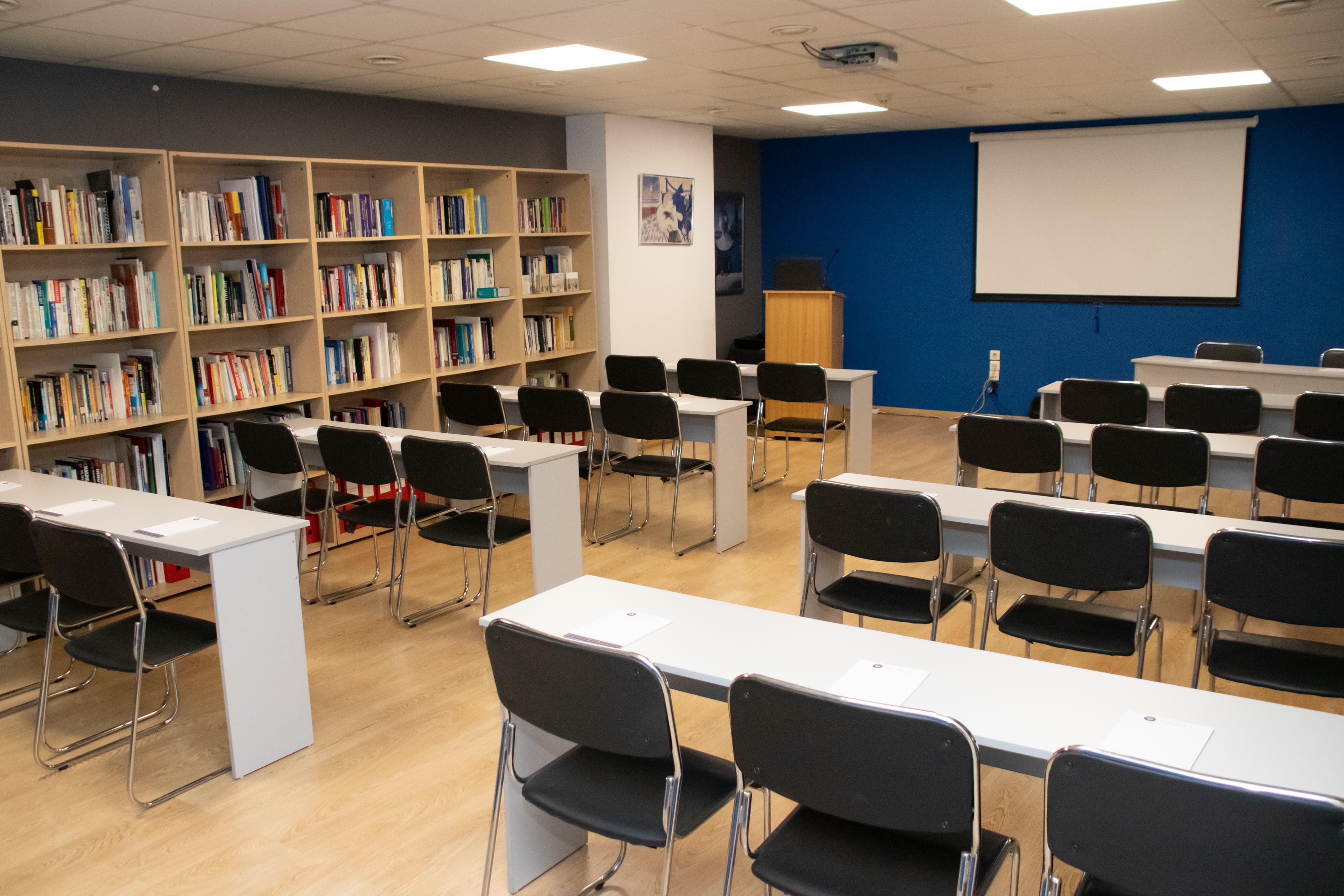 Classroom-style meeting room with rows of tables and chairs, a podium, and a presentation screen in front of a bookshelf wall.